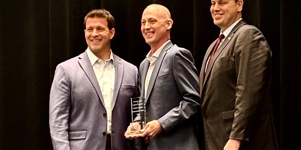 Three men in suits stand on a stage; the middle man holds an award.