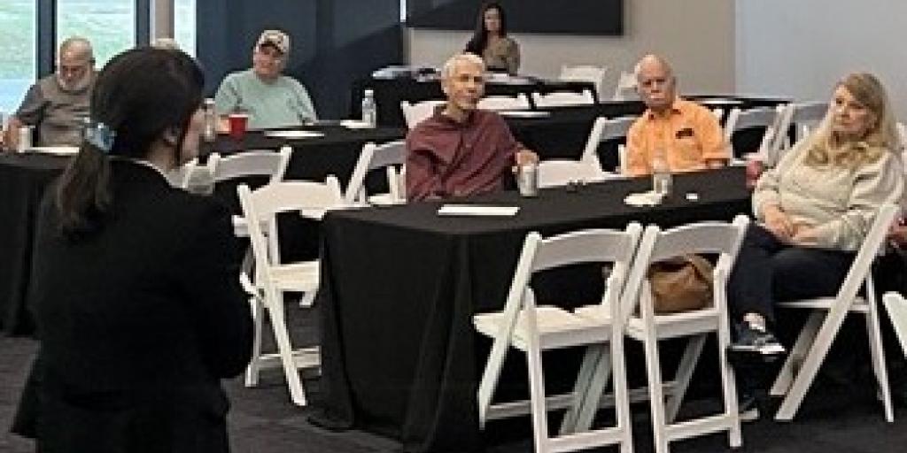 Woman in black speaks to group of people seated at tables.