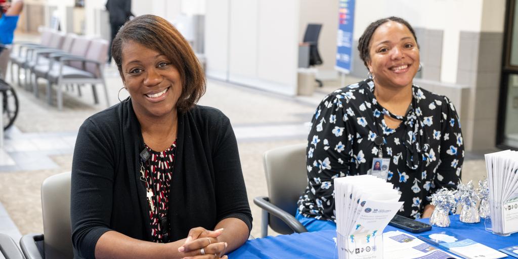 Two women sit at a table with pamphlets and baskets.