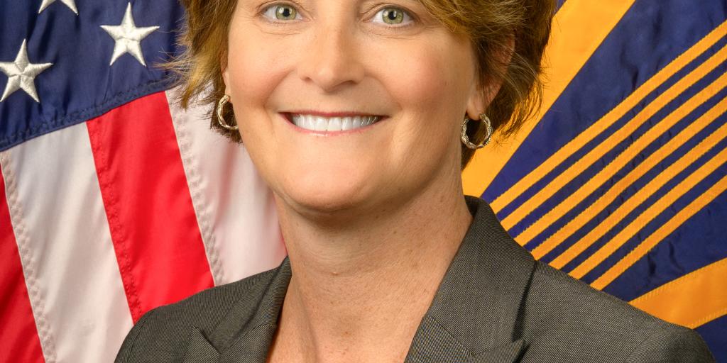 Woman in professional attire smiles in front of American and agency flags.