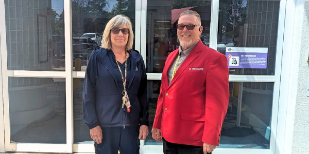 Two people stand outside a building, one in a red jacket and the other in a blue blazer.
