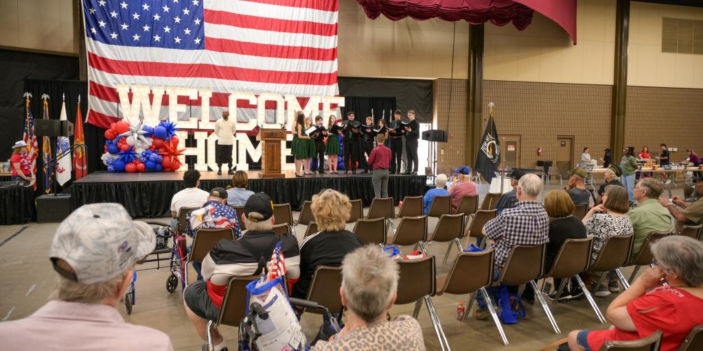 A crowd of people in a large hall with an American flag and "WELCOME HOME" banner.