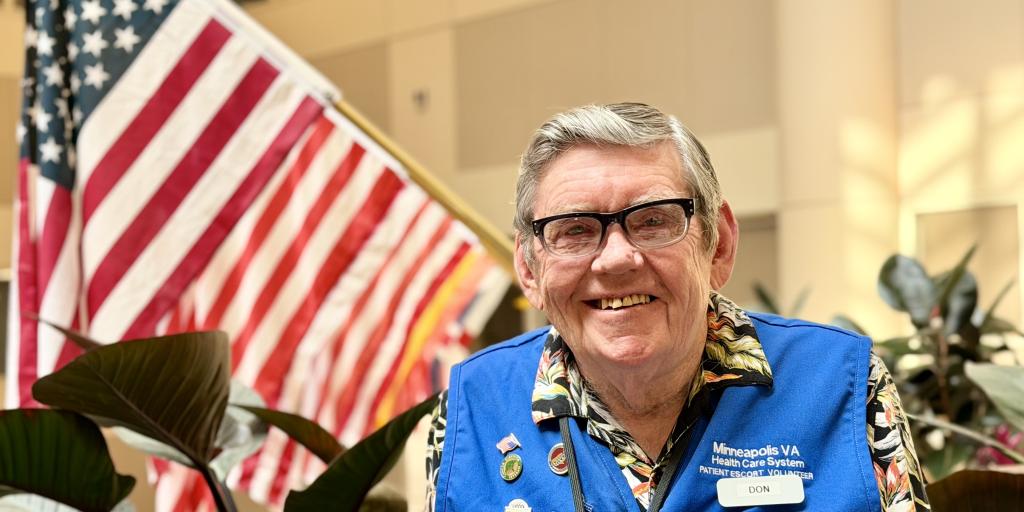 Man in blue vest and floral shirt smiling by American flag and plants.