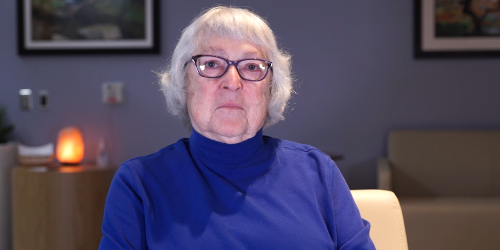 Elder woman in blue shirt and glasses sits in office.