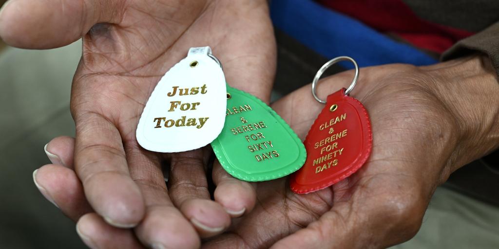Hands holding three keychains that say "Just For Today", "A Day At A Time", and "It Works If You Work It".