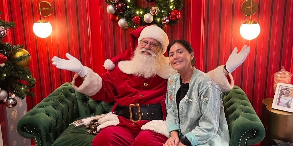 Santa Claus and a woman in a green chair with a Christmas tree and decorations in the background.