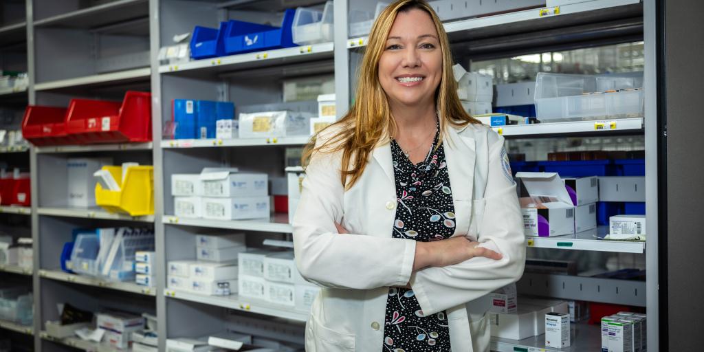 Smiling woman in white lab coat stands with arms crossed in front of shelves full of medicine.
