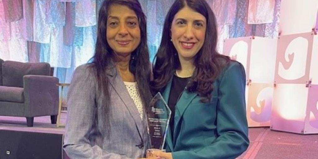 Two women, one with long hair and the other with short hair, stand on a stage holding an award.