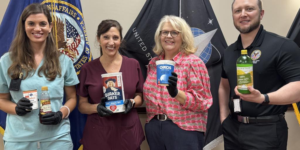 Four people in scrubs and protective gear pose with recipe ingredients in front of flags.