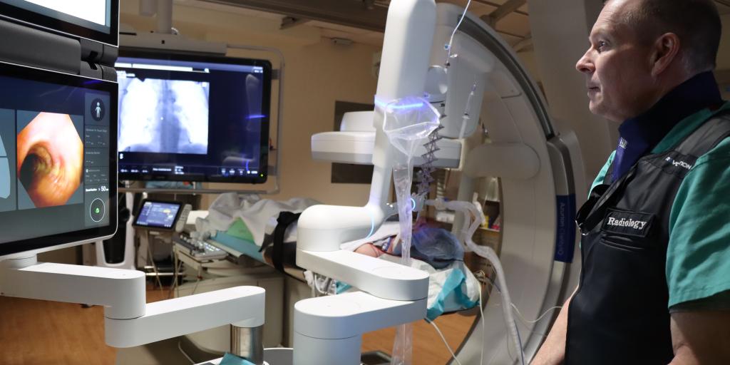 A man in green gloves and a black vest works in a medical room with screens and equipment.