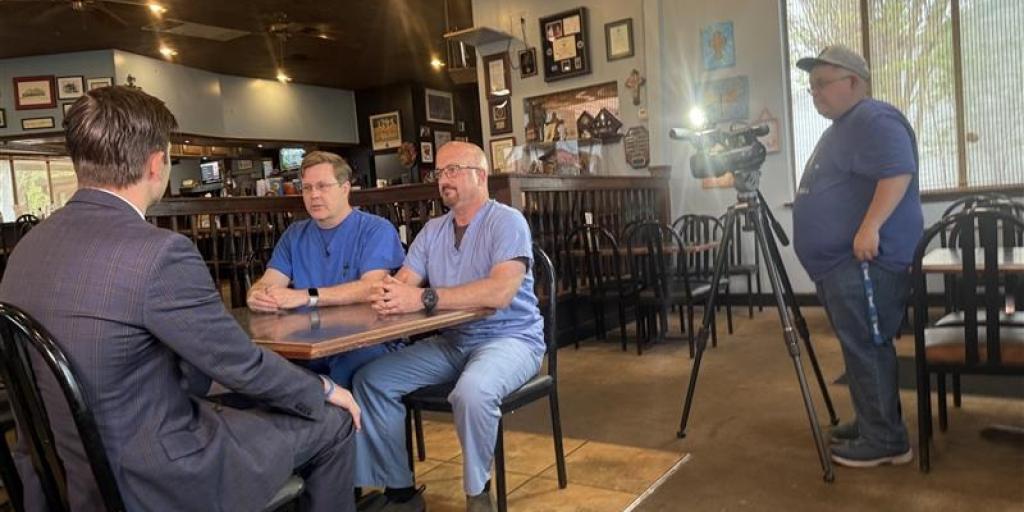 Three men in a restaurant sitting at a table with a camera man recording them.