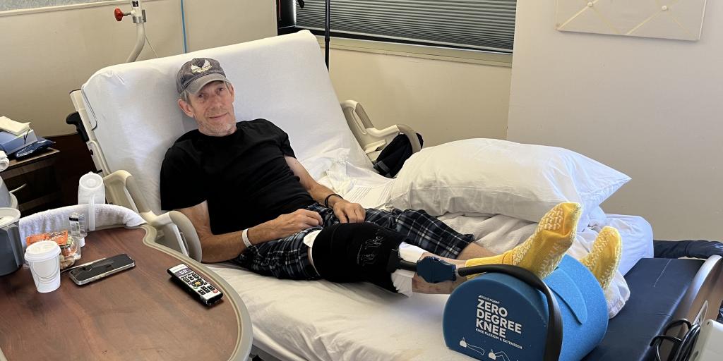 Man in hospital bed with cast on leg, lying back with remote and cup nearby.