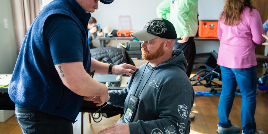 A man in a wheelchair receives assistance from another man in a blue vest.
