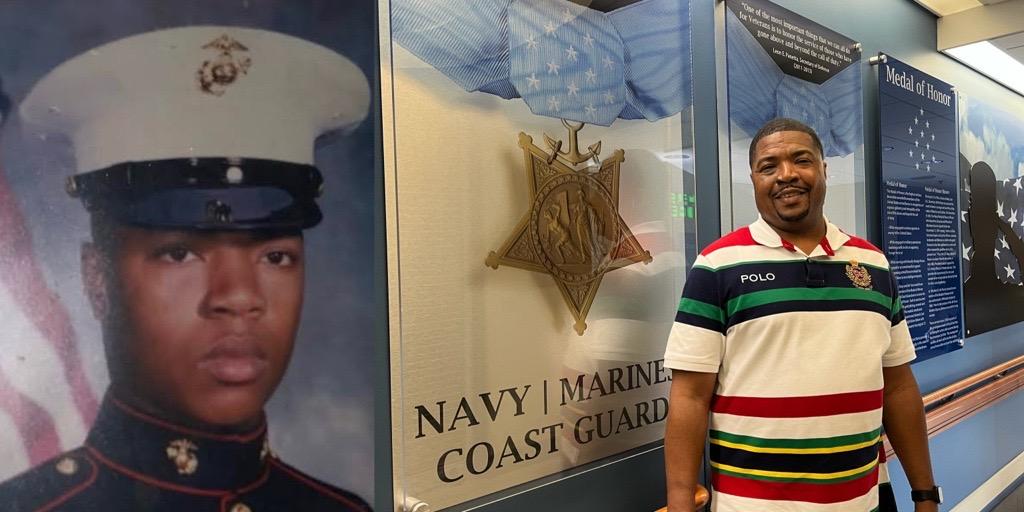 Man in striped shirt stands next to poster of Navy Coast Guard and military man.