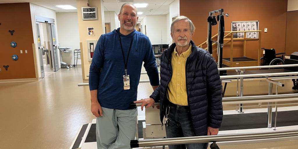 Two men in a rehab facility, one holding a handrail.