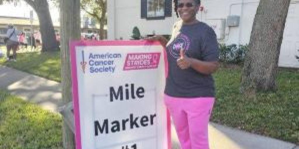 Woman in pink pants and gray t-shirt stands next to Mile Marker #1 sign.