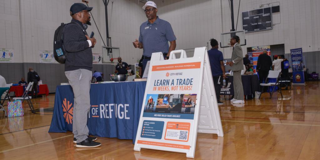Two men talking next to a "Learn a Trade" sign at a career fair in a gymnasium.