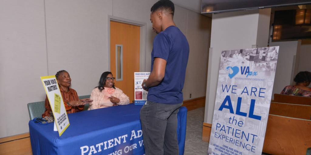 Man stands at blue table with two seated women, "We are all the patient experience" sign in background.