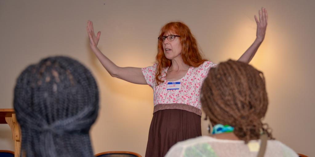 A woman with red hair and glasses stands with arms raised, speaking to a group of seated people.