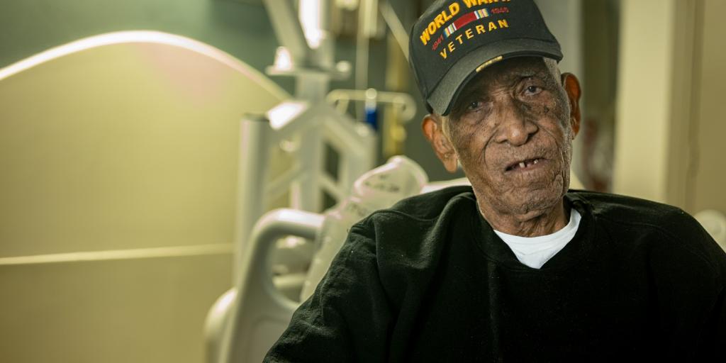 An elderly man in a World War II veteran hat sits in a hospital bed.