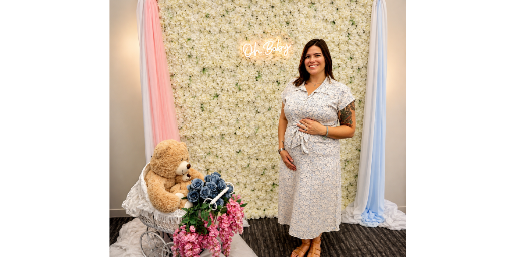 Woman with hand on belly stands in front of floral background and baby items.
