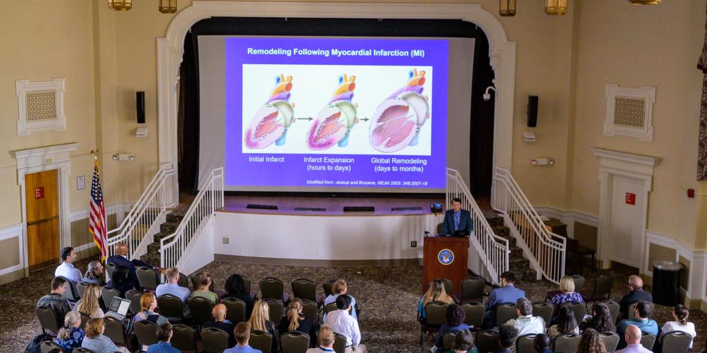 Large lecture hall with people seated facing a stage with a large screen displaying a medical diagram.