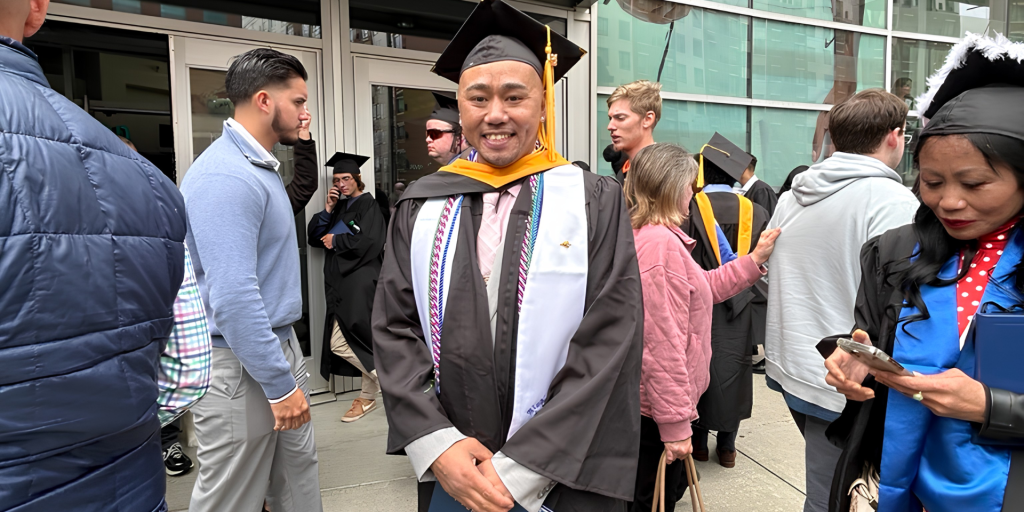 A graduate in cap and gown stands smiling among other graduates outside a building.