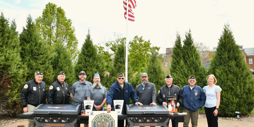 Group of people stand behind two grills at an outdoor event.