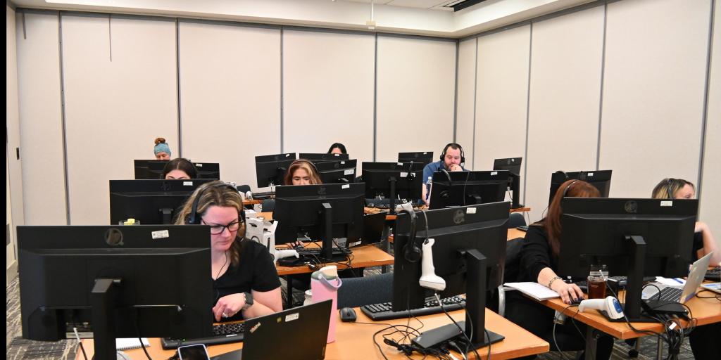 A group of people working at desks in an office with computers and laptops.