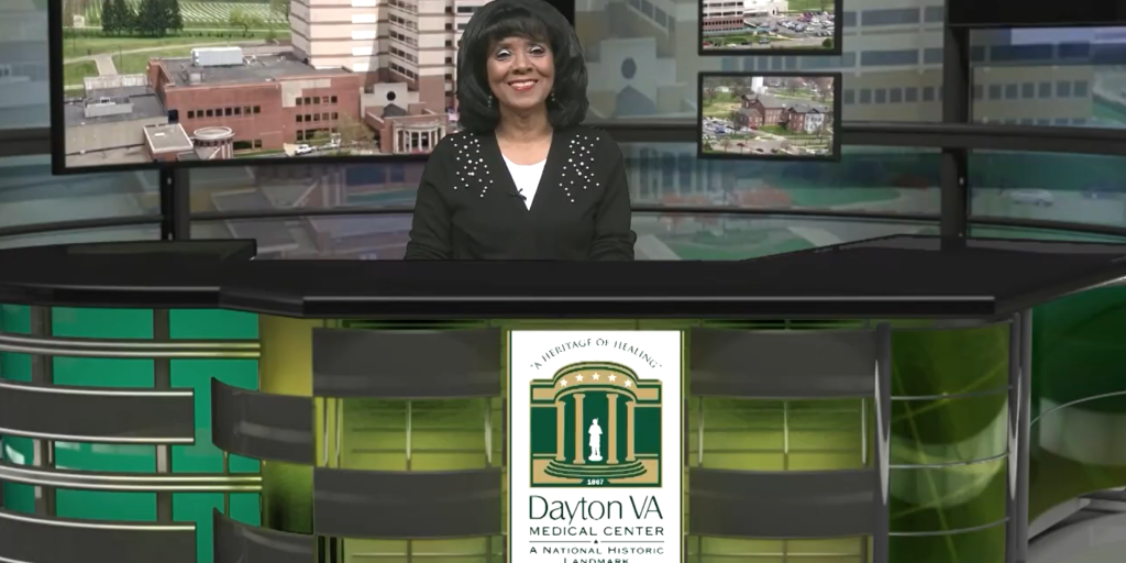 A woman stands behind a green and black news desk.