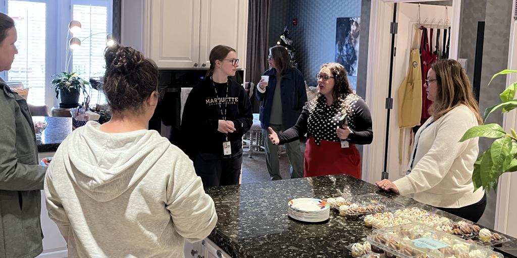 Five women chat around a kitchen island with cupcakes and coffee.