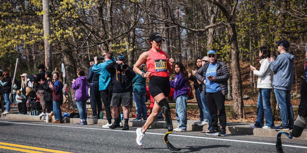 Runner with prosthetic legs crosses street at marathon, crowd watches.