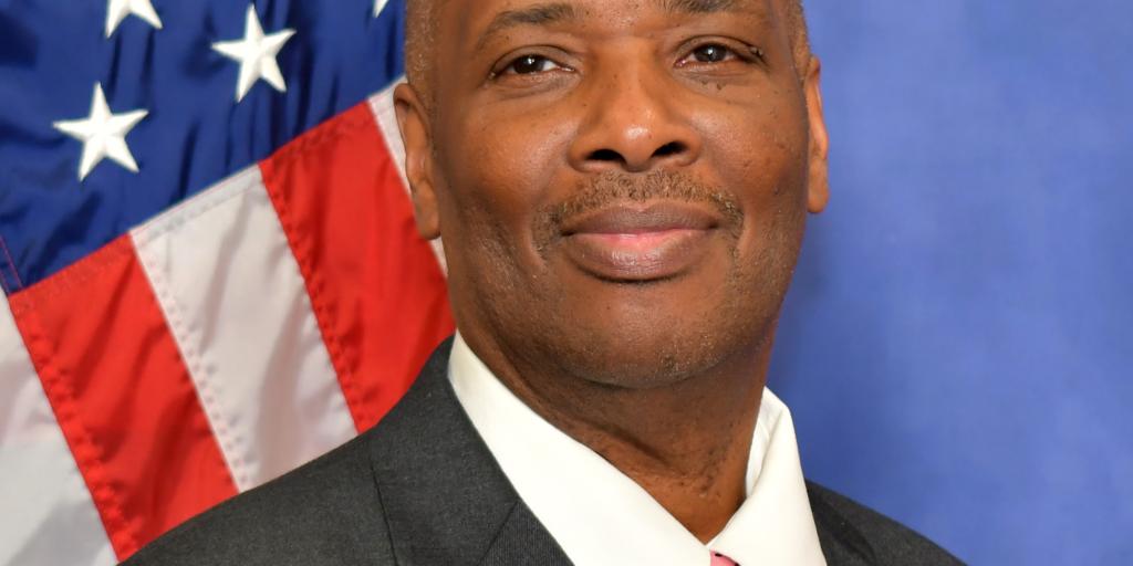 Man in suit and pink tie stands in front of American flag.