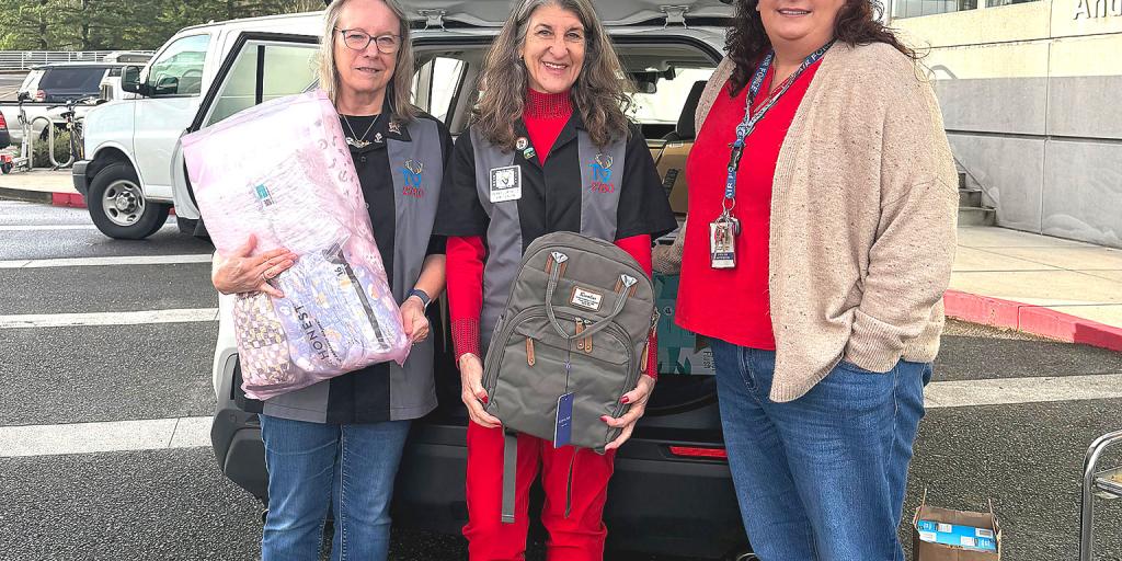 Three women in scrubs holding a mattress and backpack next to a van.