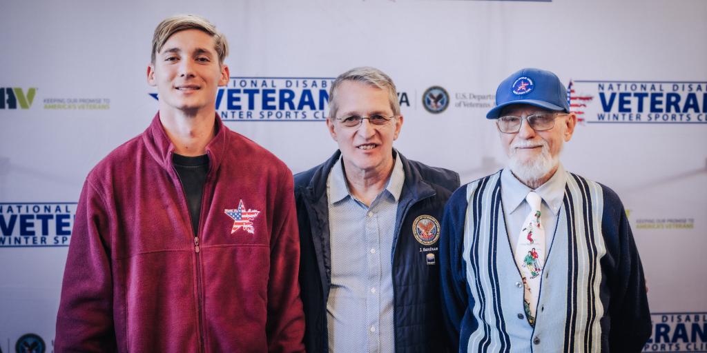 Three men standing together in front of a veterans banner.