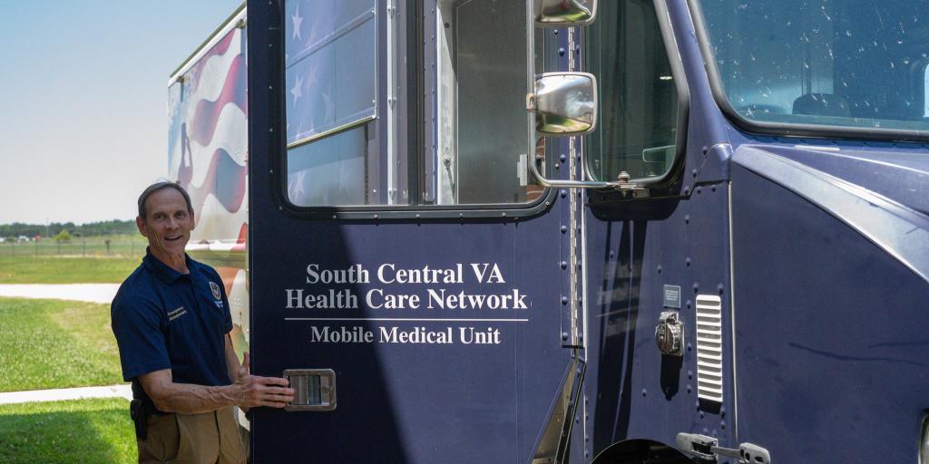 Man in blue shirt and brown pants standing by blue mobile medical unit.