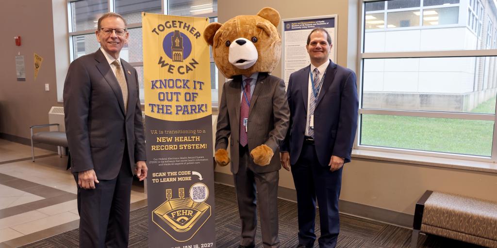Three men in suits with a bear mascot at an event.