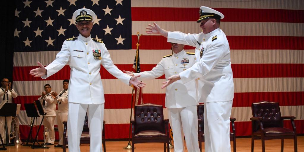 Two navy officers shake hands on stage in front of an American flag.