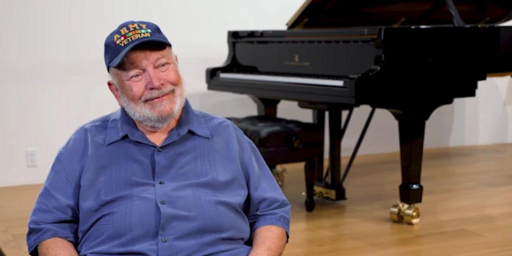 Man in blue shirt and cap sitting next to a grand piano.