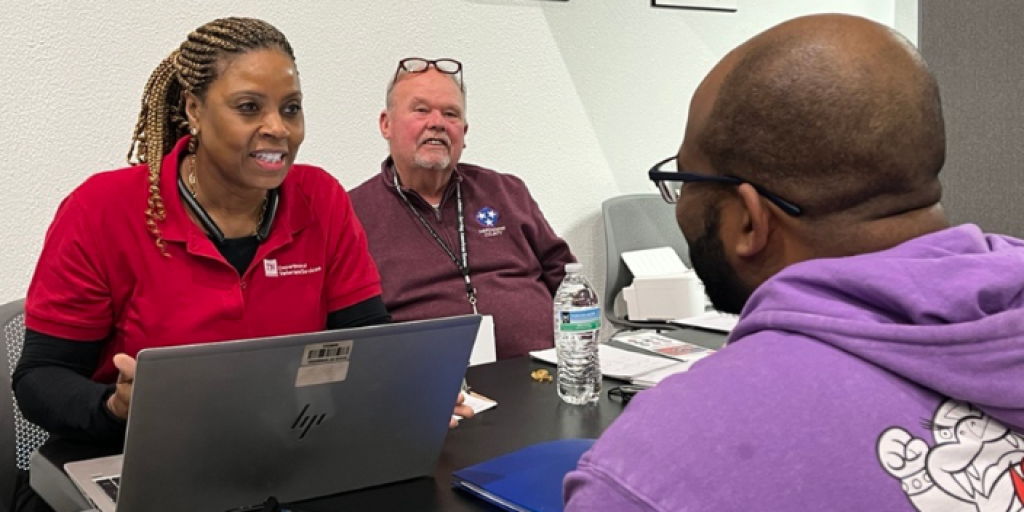 Three people at a table with a laptop, engaged in conversation.