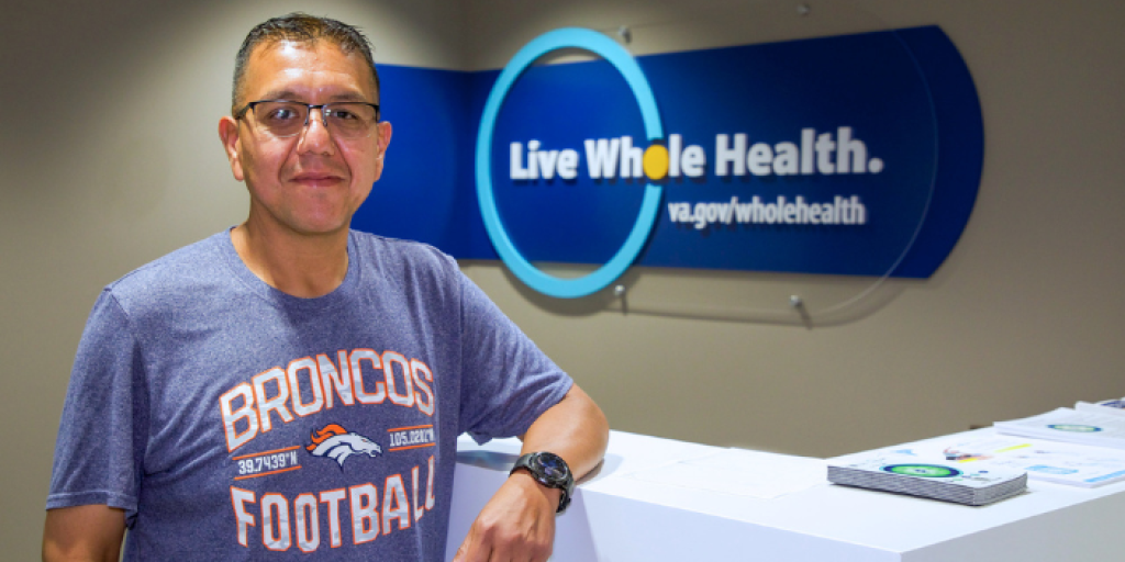 Man in blue shirt standing by desk with "Live Whole Health" sign in background.