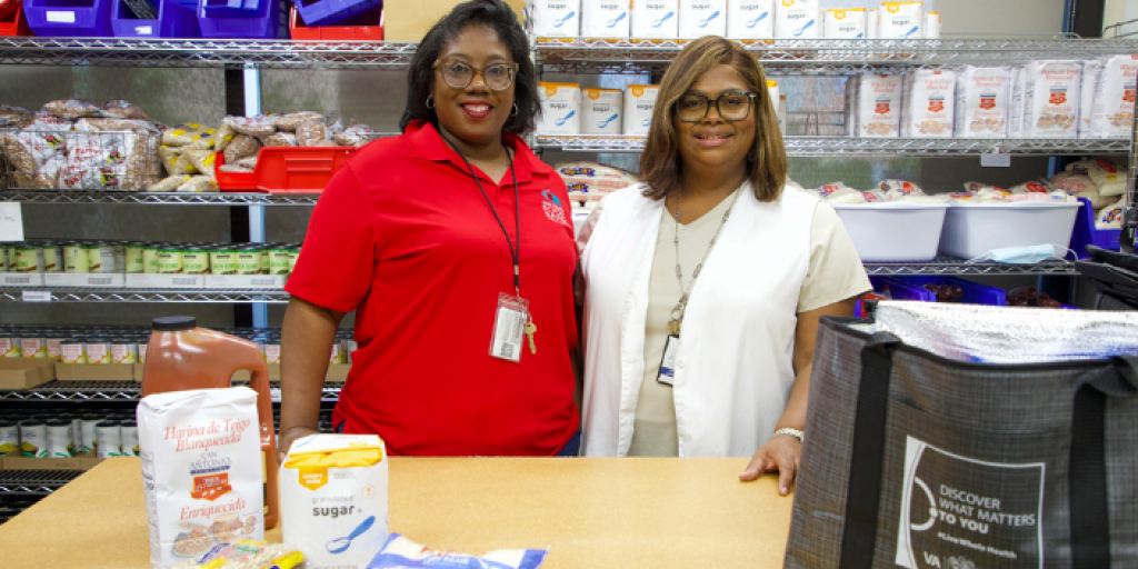 Two women stand behind a table with food items, smiling.