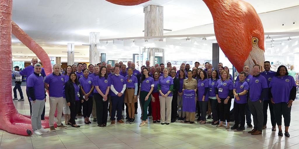 A large group of people in purple shirts pose for a photo in front of a giant pink flamingo sculpture.