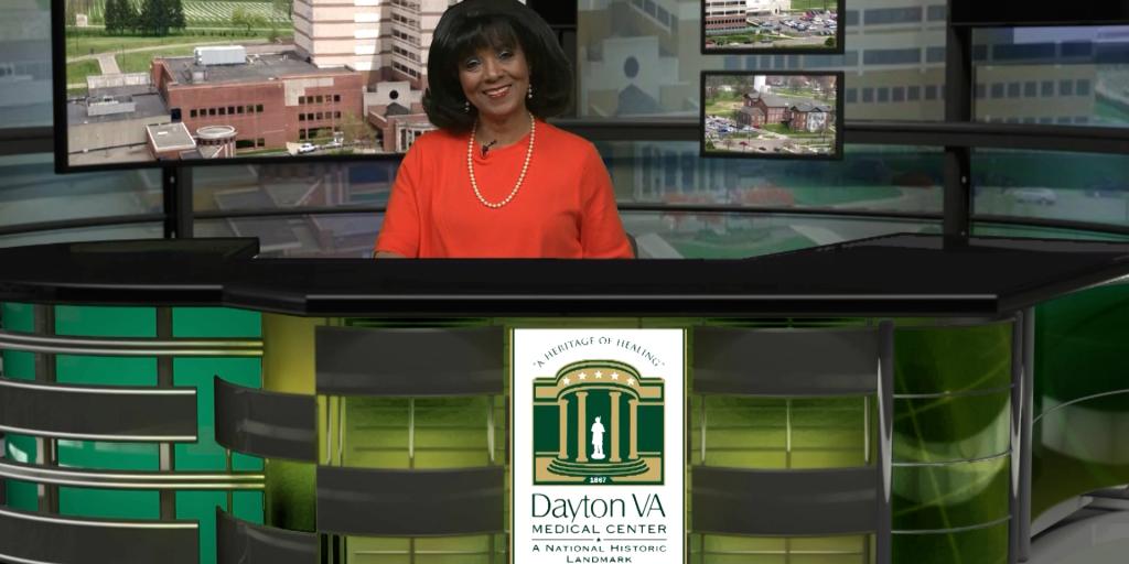 Woman in orange blouse stands behind news desk with Dayton VA Medical Center logo.