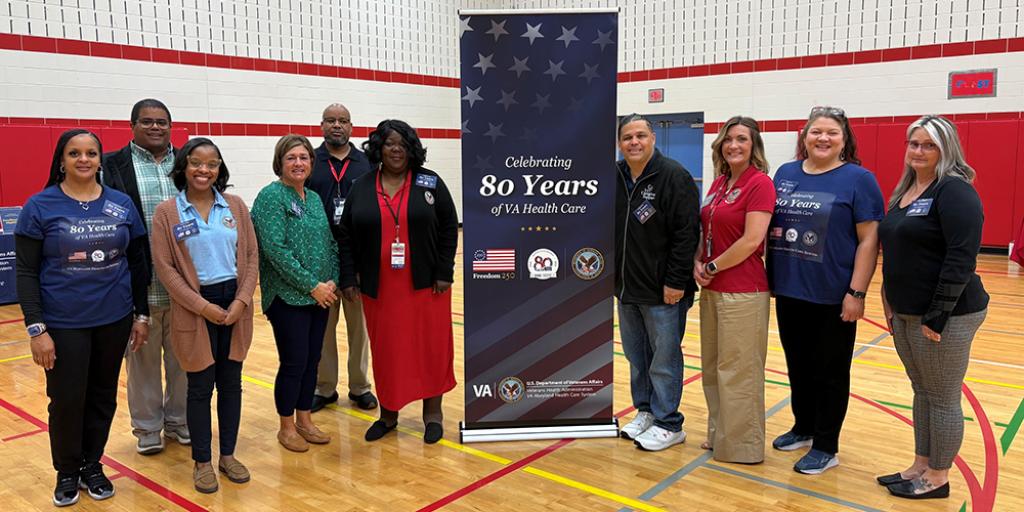Group photo of 11 people standing next to a blue banner in a gymnasium.