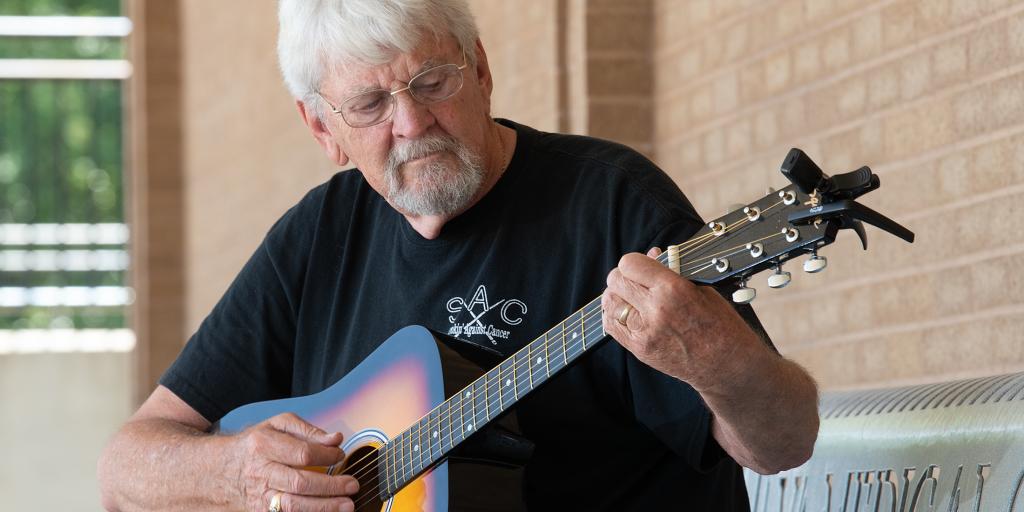 Older man with white hair and beard playing guitar outside.