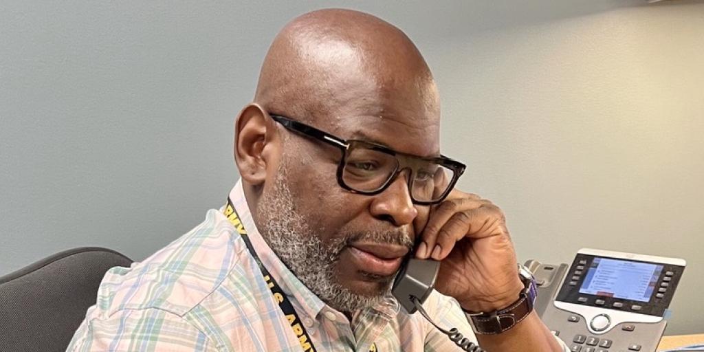 Man in plaid shirt and glasses sitting at a desk, talking on the phone.