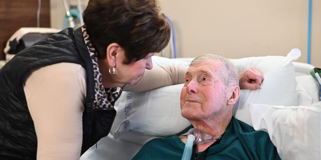 Wife leans over hospital bed to speak to elderly man with oxygen tube.