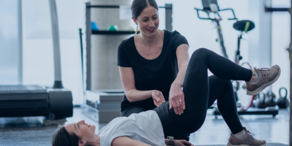 Two women in a gym, one lying on floor with raised leg, assisted by other woman.
