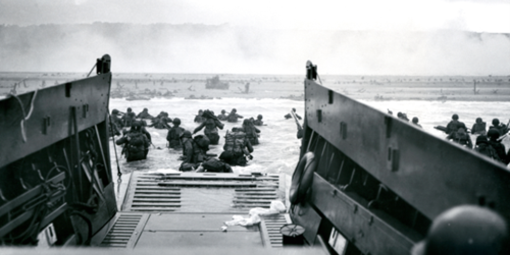 Black and white photo of soldiers disembarking from landing craft onto beach.